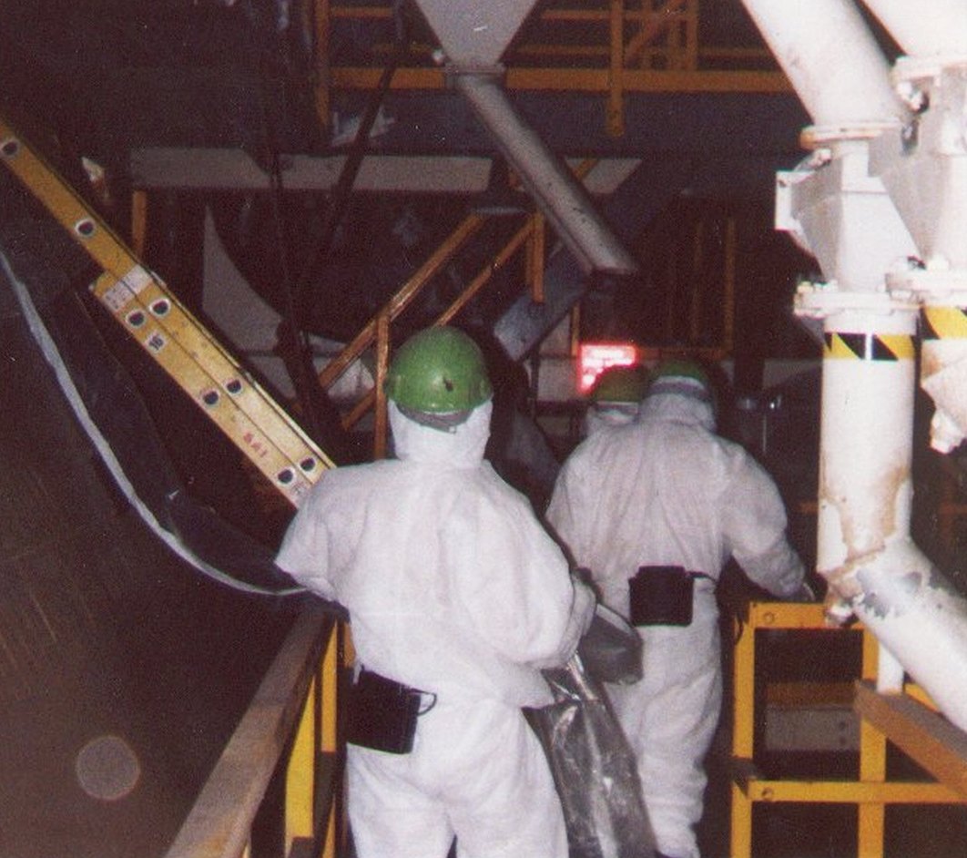 Two workers in white hazmat suits and green hard hats on a catwalk.
