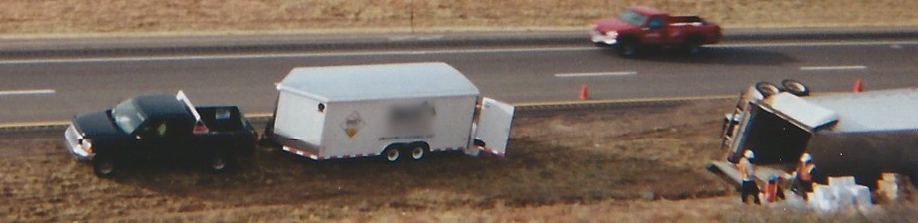 A pickup truck and hazmat trailer on a highway shoulder next to an overturned truck, with a crew at work.